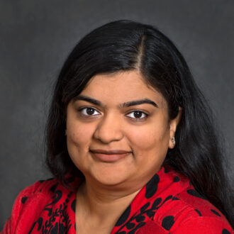 Deepti Tanjore, a person with long dark hair wearing a red top with black pattern, photographed against a gray backdrop.