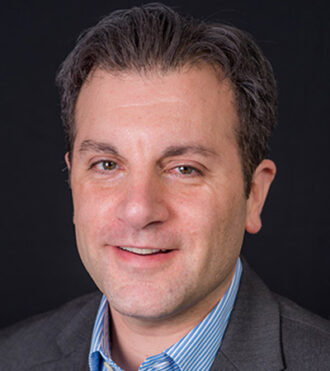 Arman Shehabi, a person with short brown hair wearing a gray blazer over a light blue and white striped collared shirt, photographed against a black background.