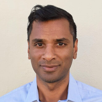 Prakash Rao, a person with short dark hair wearing a light blue collared shirt, photographed against a white wall.