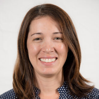 Vi Rapp, a person with medium-length brown hair wearing a navy blue and white polka-dotted top, photographed against a white background.