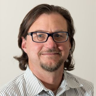 David Alumbaugh, a person with bobbed length brown hair wearing glasses and a white collared shirt with gray stripes, photographed against a beige wall.