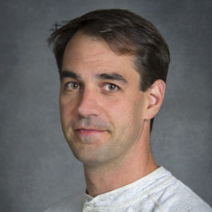 Greg Hura, a person with short brown hair wearing a white top photographed against a gray backdrop.