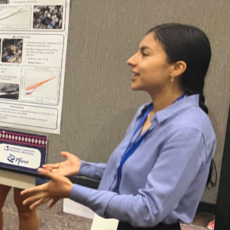 A person with long dark hair pulled back wearing a blue collared shirt talks about their research next to their poster board at a conference.