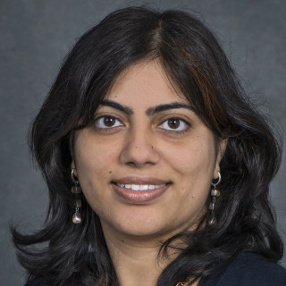 Bhavna Arora, a person with medium-length dark hair wearing a dark top with dangling earrings, photographed against a dark gray backdrop.