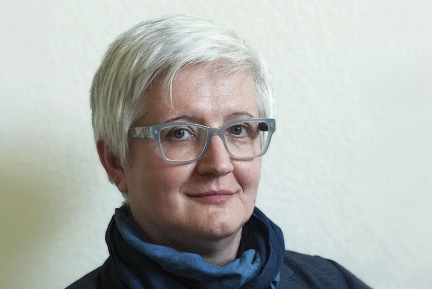 Person with short white hair wearing glasses and a blue scarf.