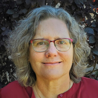 Lori Zscherpel, a person with medium-length gray hair wearing red glasses and a red top, photographed outdoors.