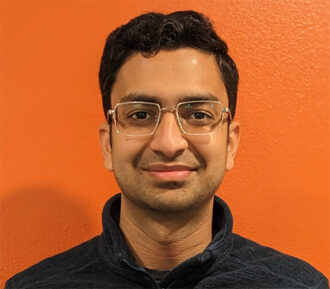 Mukul Dave, a person with short black hair with glasses wearing a dark jacket against an orange background.