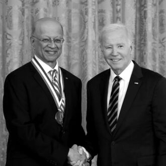 Two people in dark suits and ties shaking hands. The individual on the left is wearing a National Medal of Technology and Innovation.