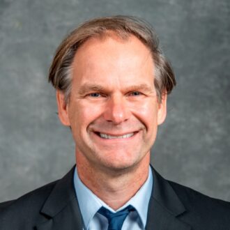 Thomas Schenkel, a person with short gray hair wearing a suit and posing for a portrait. Photographed against a gray backdrop.