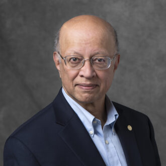 Portrait of Ashok Gadgil, a person wearing glasses and a black blazer with light blue collared shirt. Photographed against a gray backdrop.