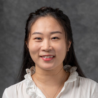 Portrait of Zhi (Jackie) Yao, a person with long dark hair wearing a white blouse. Photographed in front of a gray backdrop.