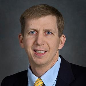 David Schlegel, a person with short brown hair, wearing a black blazer over a light blue collared shirt and yellow and blue striped tie. Photographed against a gray backdrop.