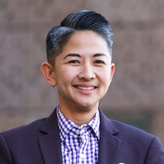 Lady Idos, a person with short gray black hair wearing a dark purple blazer over a white and purple striped collared shirt.