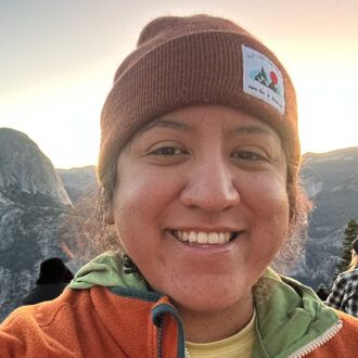 Sonia Vargas, a person wearing a brown beanie and orange fleece jacket, smiling for a selfie outdoors.