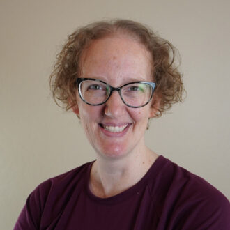 Tracy Mattox, a person with short curly light brown hair wearing classes and a mauve shirt, photographed against a light colored background.