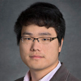 Qing Zhu, a person with short dark hair wearing glasses and a dark blazer over a light pink collared shirt. Photographed against a gray backdrop.