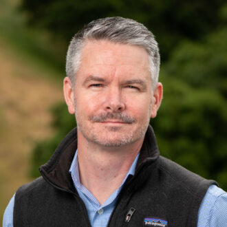 Eoin Brodie, a person with short gray hair wearing a black Patagonia vest over a blue and white checkered collared shirt, photographed outdoors.