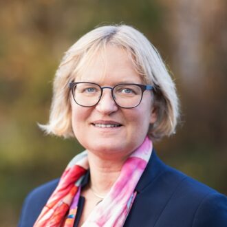 A person with short blonde hair wearing glasses and a navy coat with a colorful scarf. Photographed outdoors.