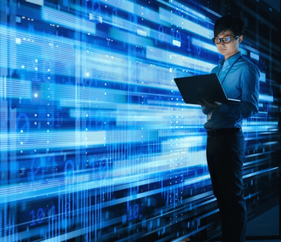 A person with glasses stands holding a laptop next to a large digital screen featuring data and graphs in a blue, modern, high-tech environment.