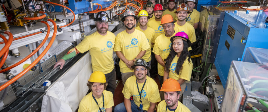 A group of people in yellow tshirts and multicolored hard hats. They are posing in a scientific facility.