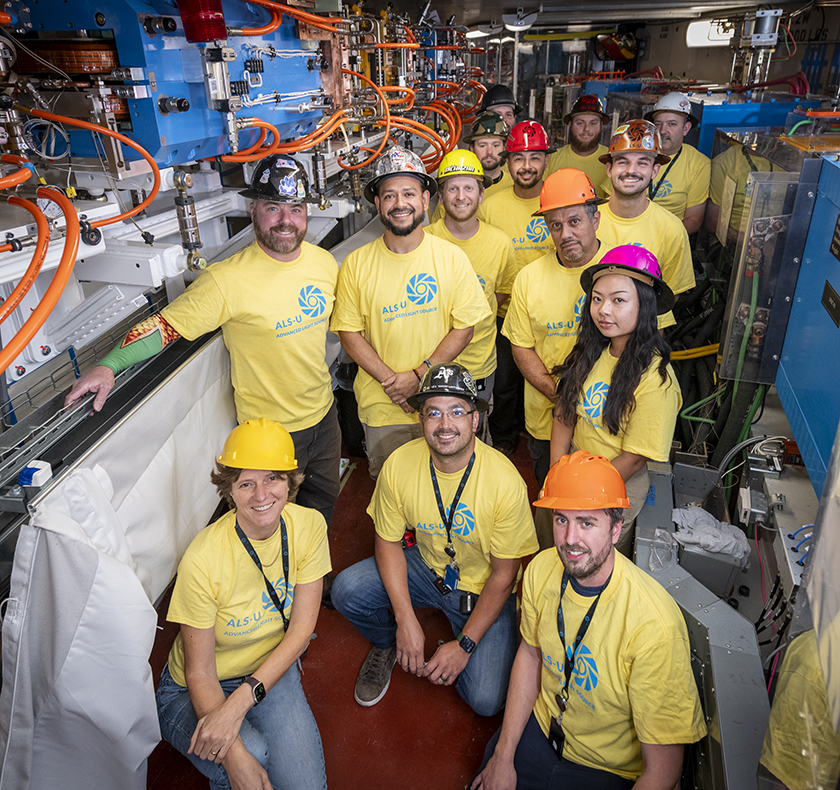 A group of people in yellow tshirts and multicolored hard hats. They are posing in a scientific facility.