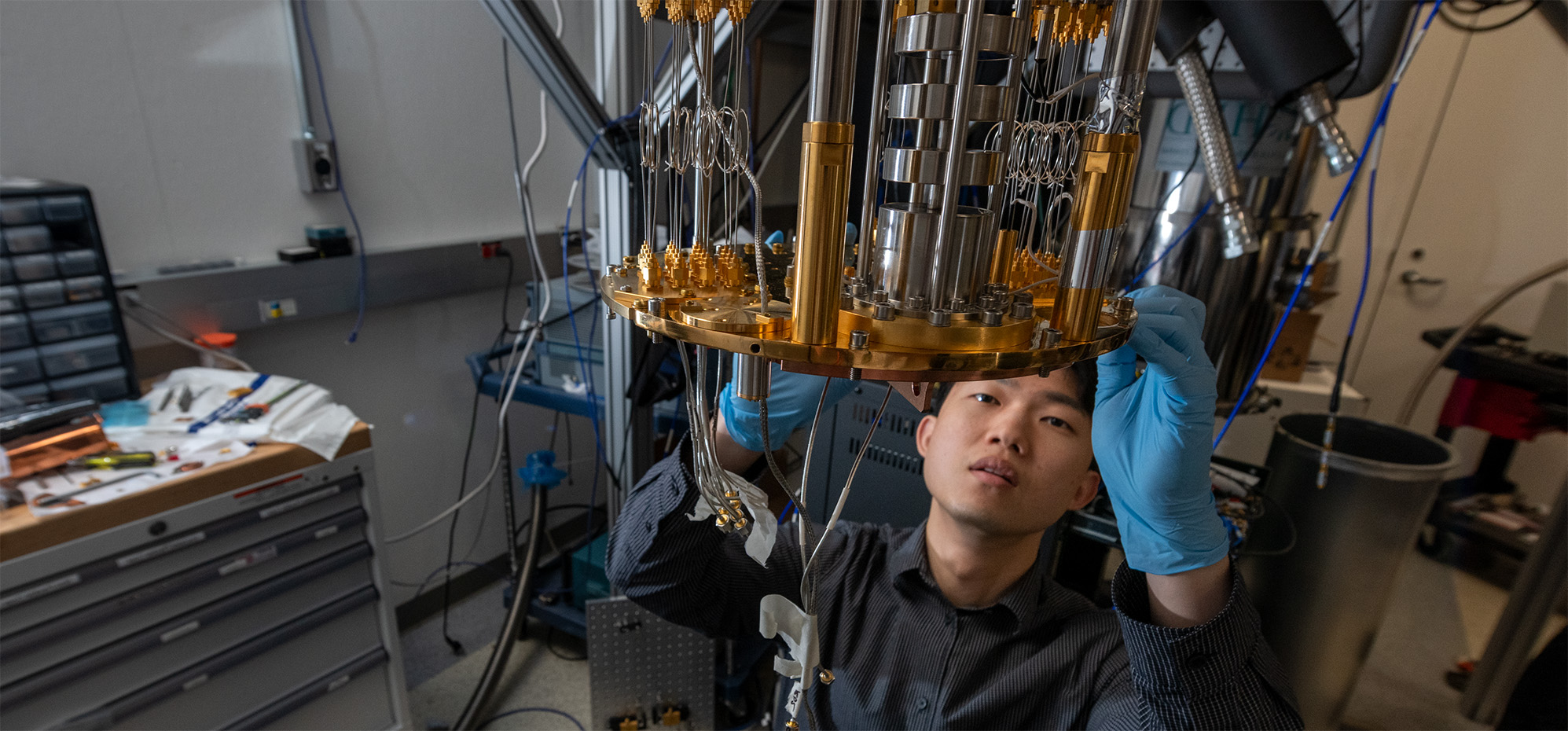 A researcher in a lab crouched below a silver and gold quantum computer. They are making adjustments to the bottom edge.