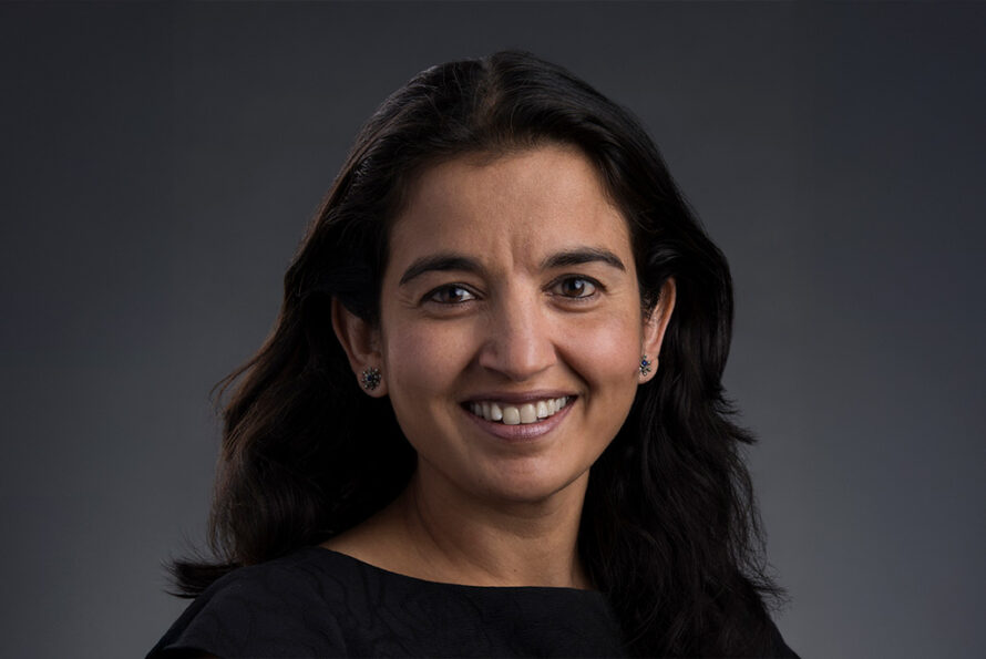 Person with medium-length dark hair wearing a black top against a gray background, smiling.