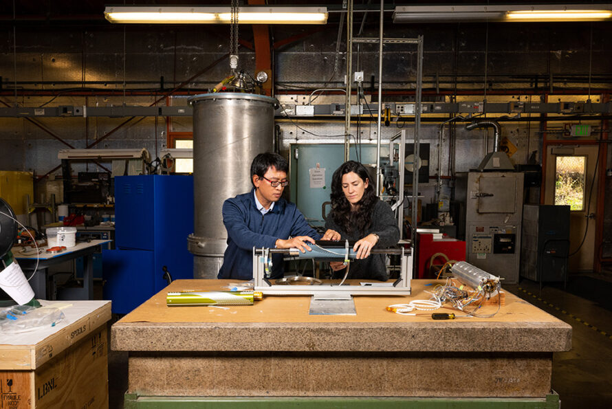Two researchers examine a cylindrical component mounted in a testing rig on a workbench in a lab filled with equipment and tools.