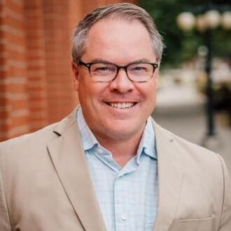 Peter Larsen, a person with short gray hair wearing dark-rimmed glasses, a collared blue checkered shirt under a tan blazer, smiling.