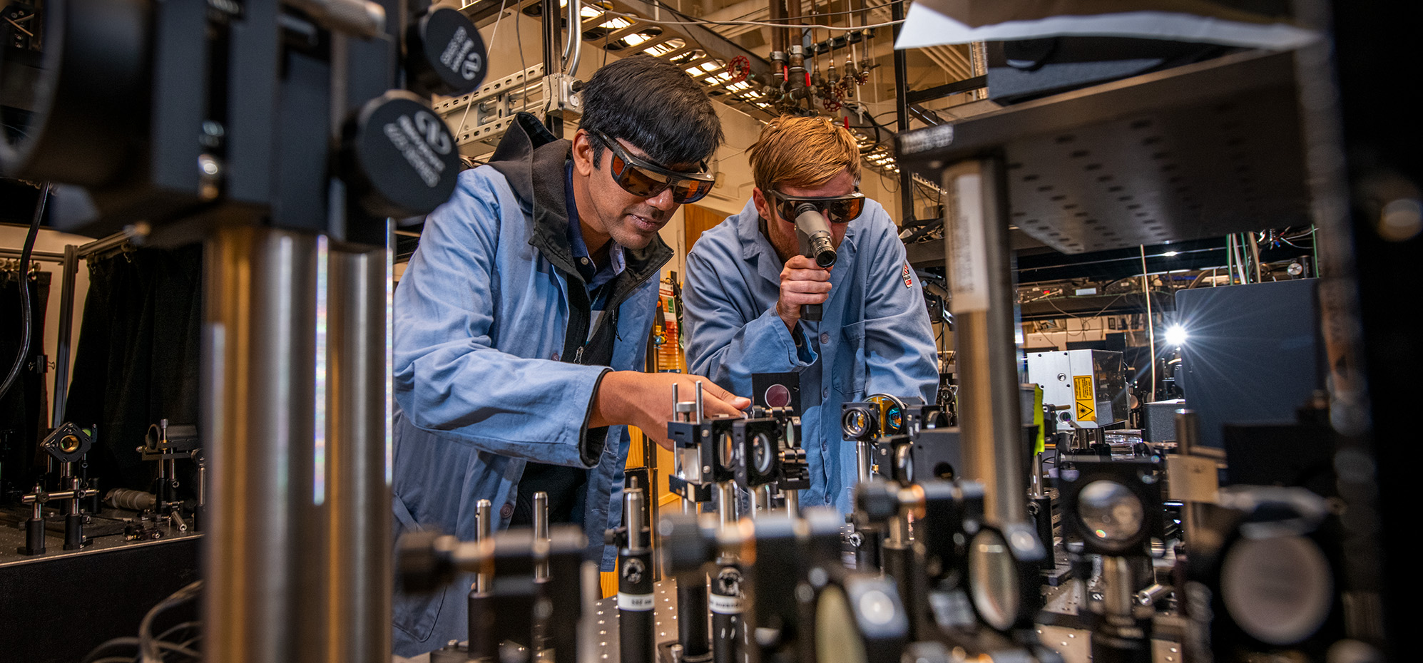Two researchers in protective glasses and blue lab coats work on a machine.