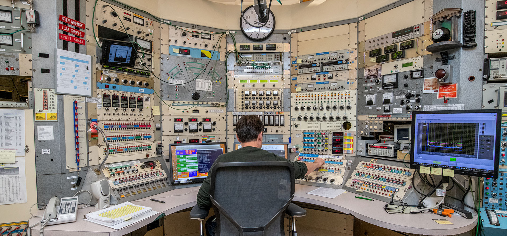 A person adjusts a setting at a console with multiple panels of knobs, switches, and screens.