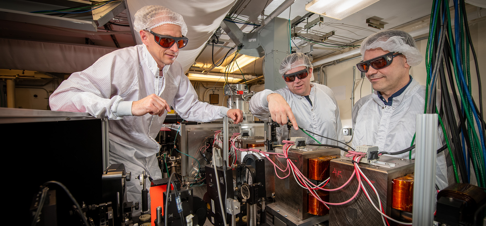Three researchers in lab coats and hairnets working on a complex machine with wires and equipment, wearing protective orange-tinted glasses.