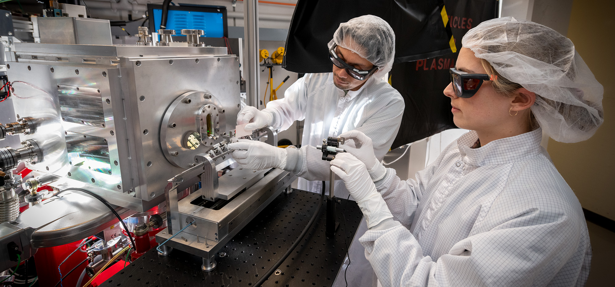 Two researchers in lab coats and hairnets working on a complex machine with wires and equipment, wearing protective glasses.