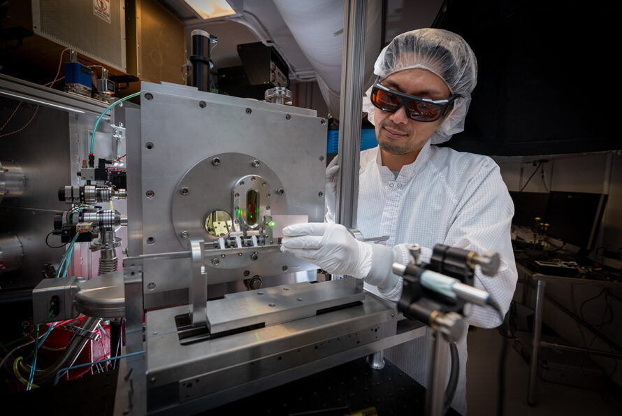 A rseearcher in a lab coats and hairnet working on a complex machine with wires and equipment.