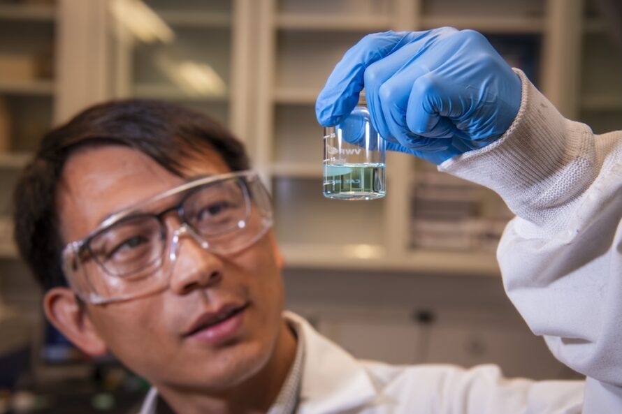 A close-up photograph of a man wearing glasses, a labcoat, and gloves who is holding up a small clear vessel containing light blue liquid.