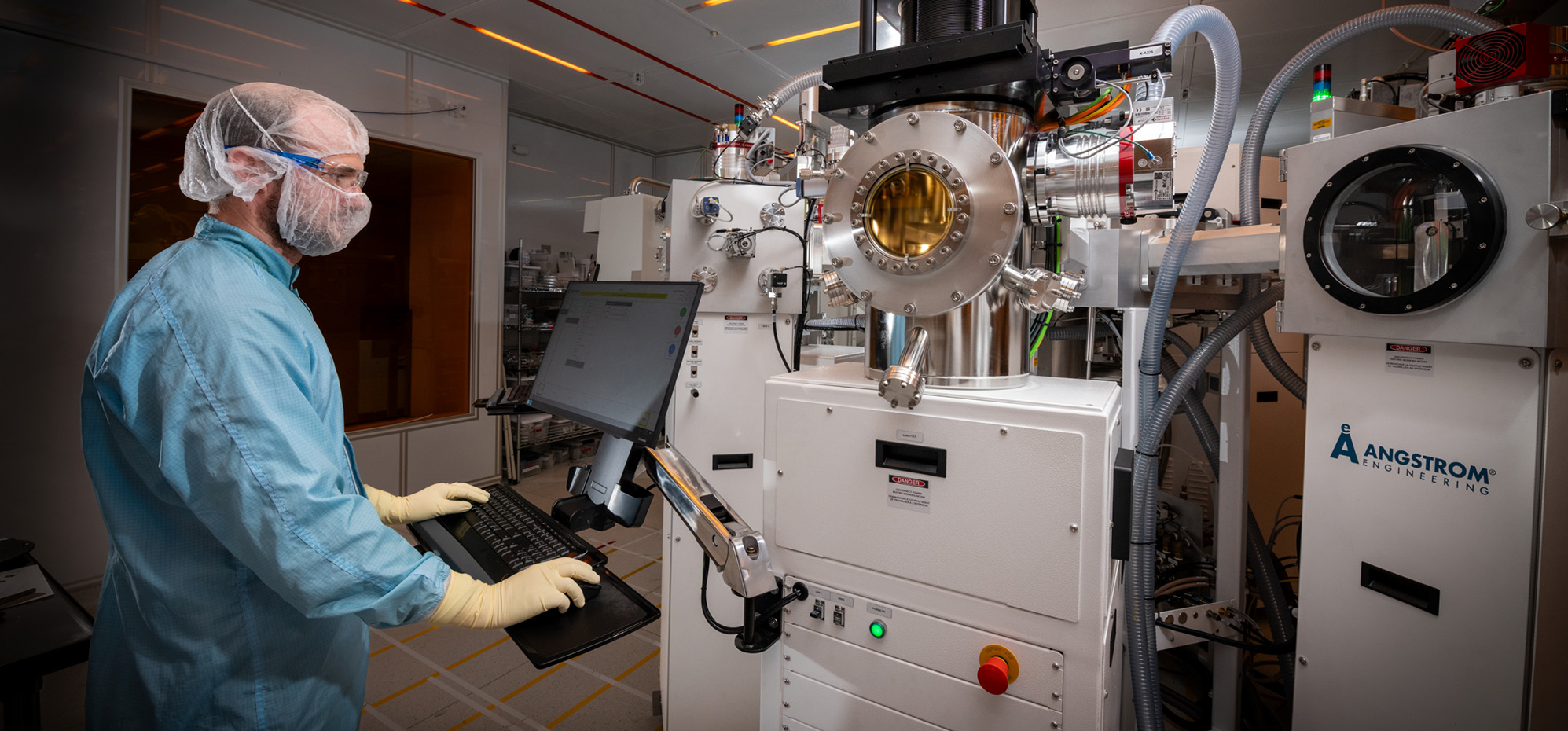 A researcher in light blue personal protective equipment works at a computer connected to the cluster tool.