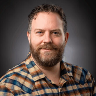 Harry Lisabeth, a person with short brown hair and a beard wearing a checkered yellow, blue, and red collared shirt, smiling for a portrait against a gradient gray background.