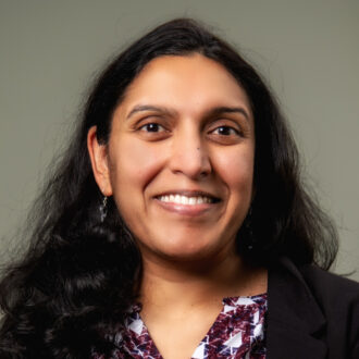 Person with long dark hair wearing a purple, blue, and white patterned shirt under a black jacket, smiling for a portrait.