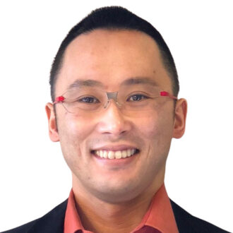 Person with short black hair wearing glasses and a red collared shirt under a black jacket, smiling in front of a white background.