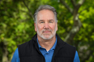 Person wearing a blue collared shirt and black vest in front of green foliage.