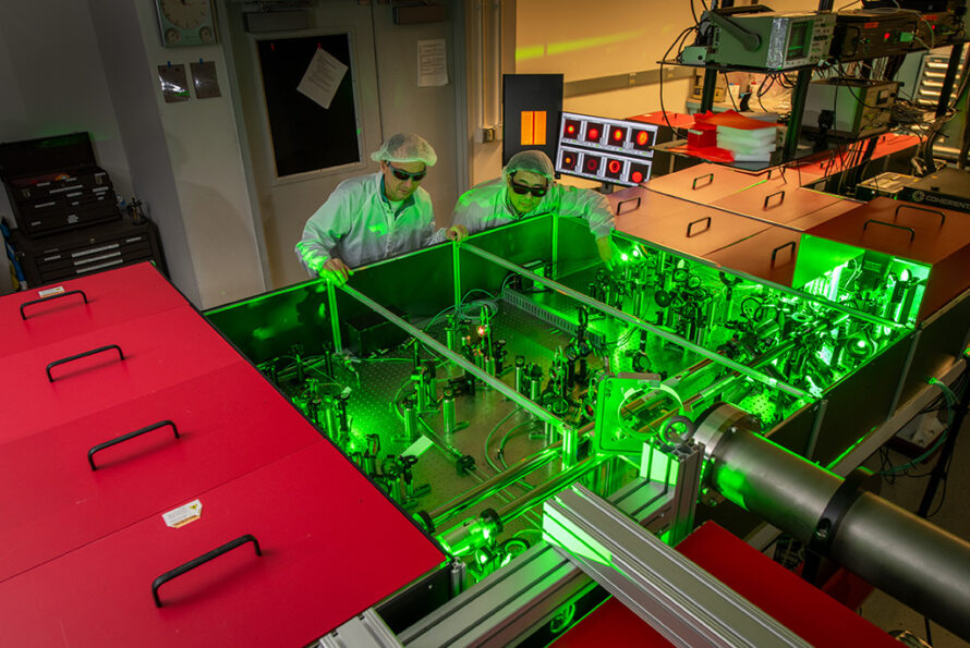 Two researchers wearing lab coats and safety glasses look at experimental equipment bathed in green light.
