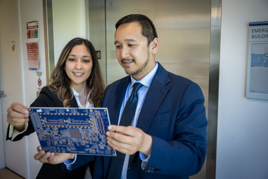 Two researchers in suits hold a device that resembles a circuit board.