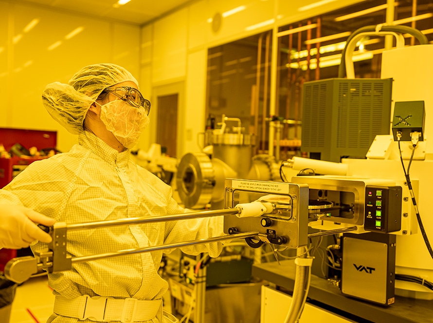 A researcher dressed in personal protective equipment operates a machine in a yellow-tinted room.