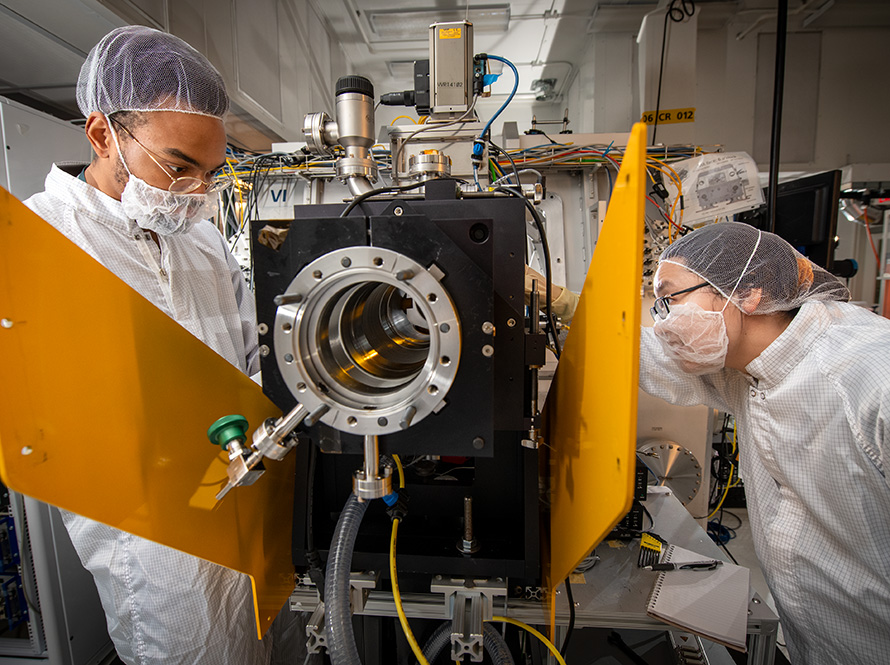 Two researchers dressed in personal protective equipment operates a machine.