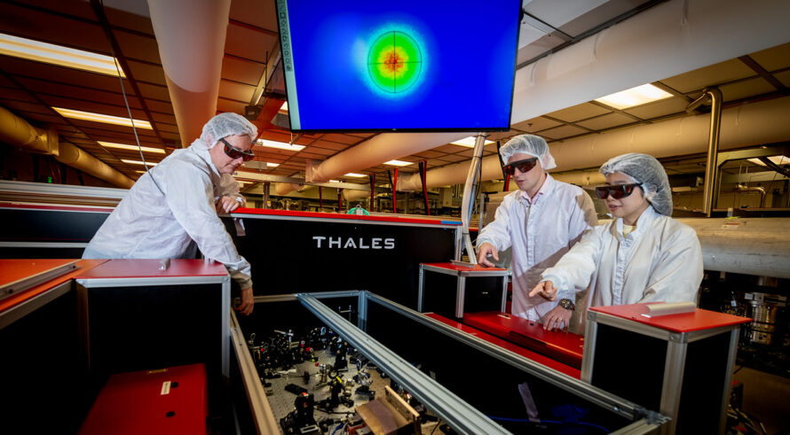 Three researchers in white coveralls and protective eyewear stand over equipment in a lab. A monitor with colorful figures is in the background.