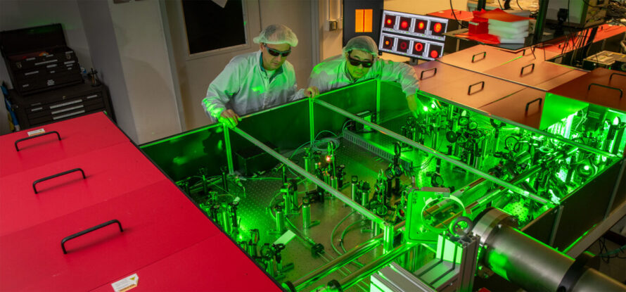 Two researchers wearing lab coats and safety glasses look at experimental equipment bathed in green light.