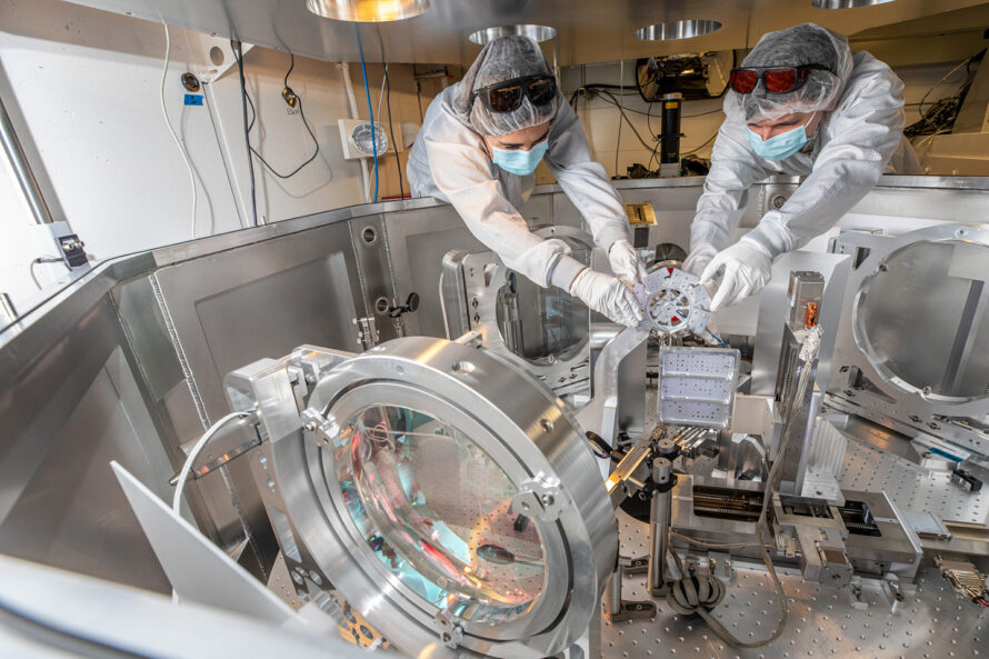 Two people in cleanroom suits install a small silver device in a small enclosure with lenses and other equipment.