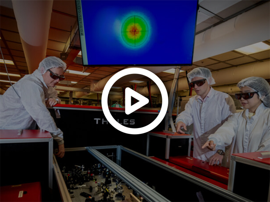 Three researchers in white coveralls and protective eyewear stand over equipment in a lab. A monitor with colorful figures is in the background.