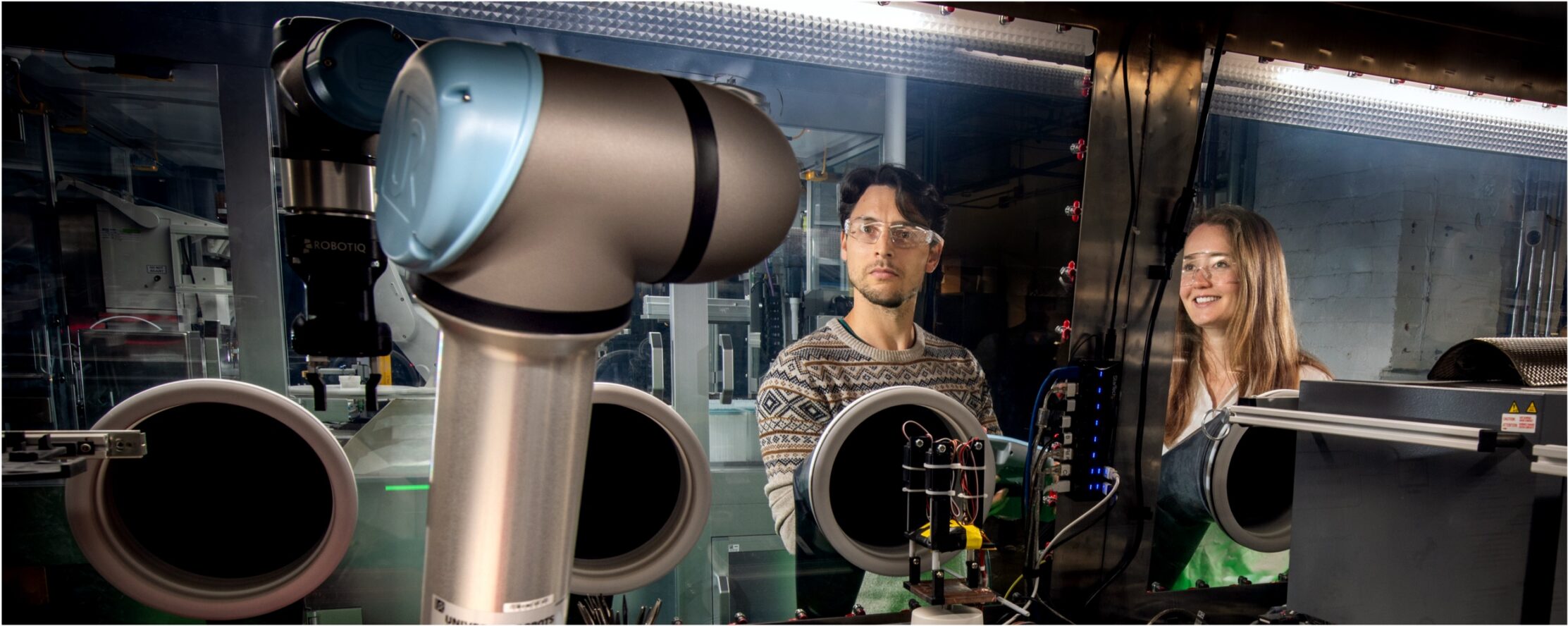 Two researchers wearing safety glasses observe an automated robotic system operating inside a laboratory equipped with enclosed instrumentation and advanced scientific equipment