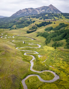 A long, windy portion of a river flowing through a blooming green mountainous landscape with clear skies